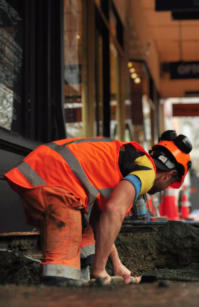 Worker in an orange safety vest and hard hat digging into a trench on a city footpath, surrounded by construction equipment and cones.