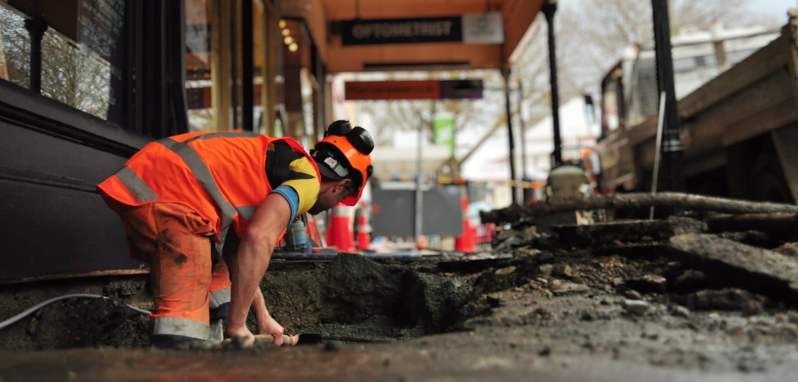 Worker in high-visibility gear kneeling and digging into a trench on a city footpath during construction work, with safety cones and shopfronts in the background.