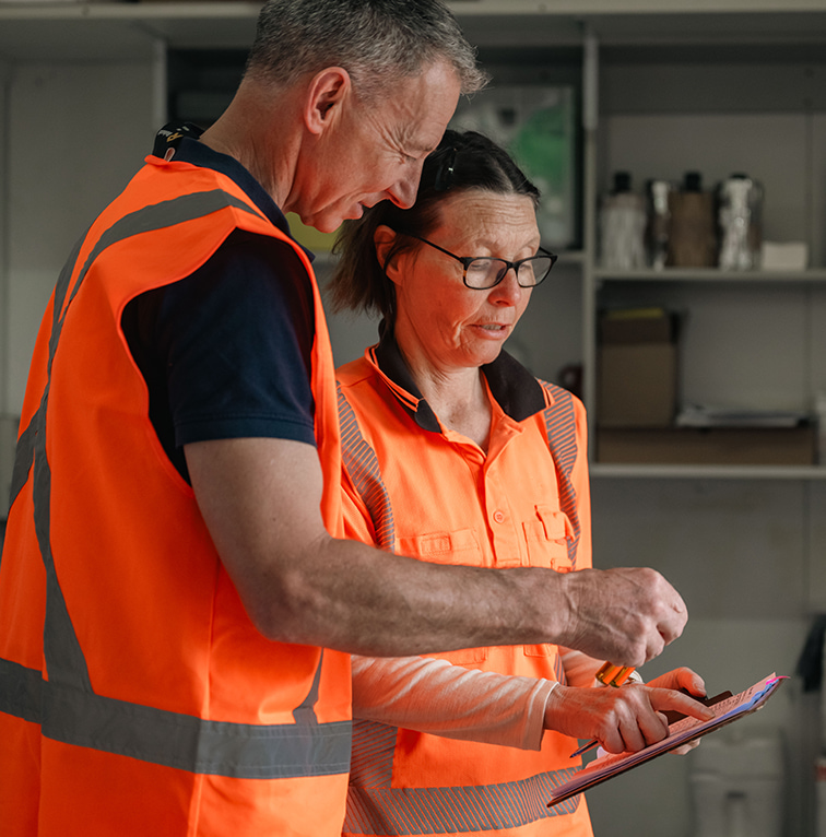 Two workers in orange high-visibility vests reviewing notes on a clipboard inside a worksite office.