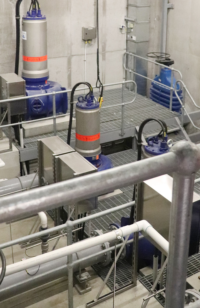 Close-up view inside an industrial pump station showing metal piping, blue pumps, and grated walkways surrounded by concrete walls and safety railings.