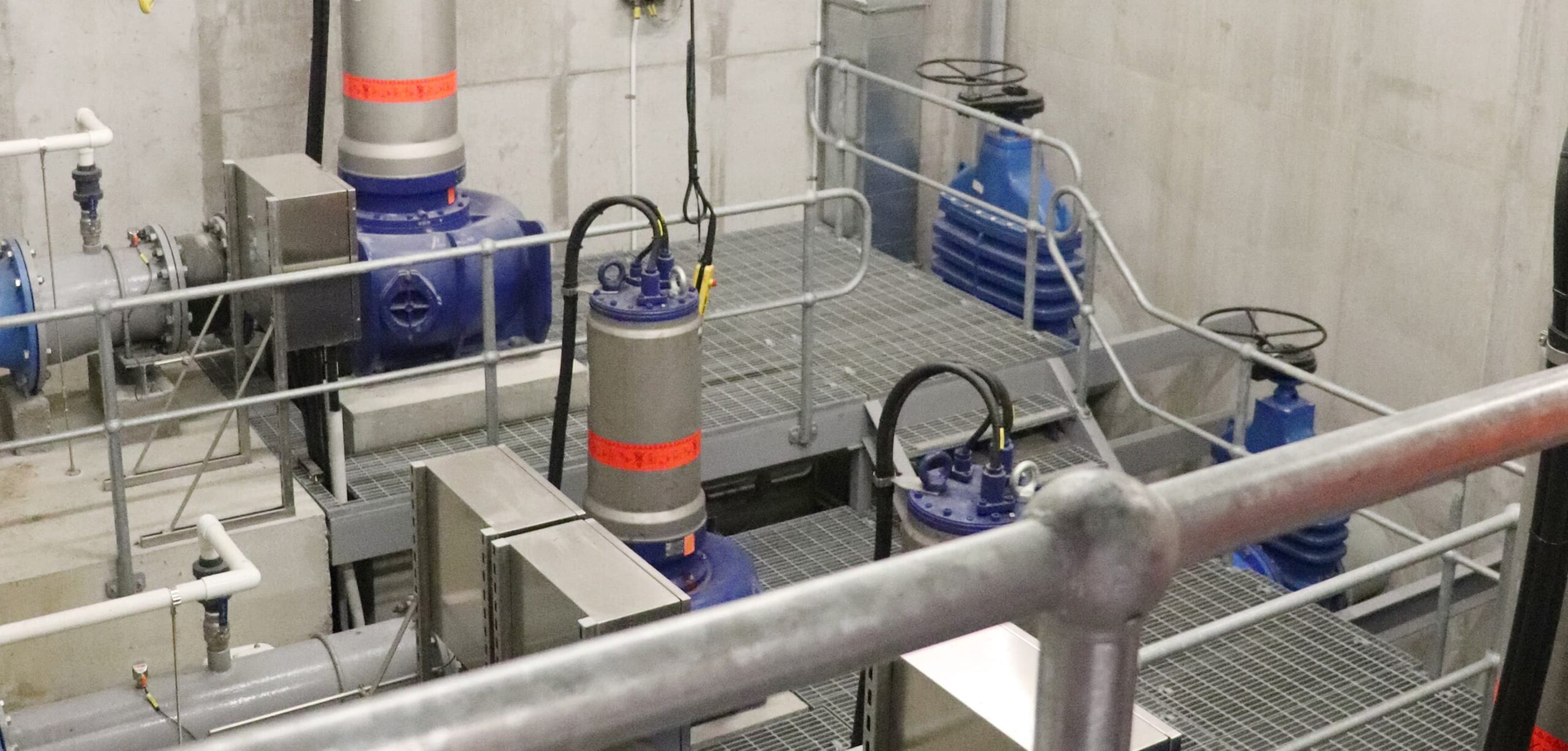 Industrial pump station interior featuring large blue pumps, metal walkways, railings, and interconnected pipes and valves inside a concrete facility.