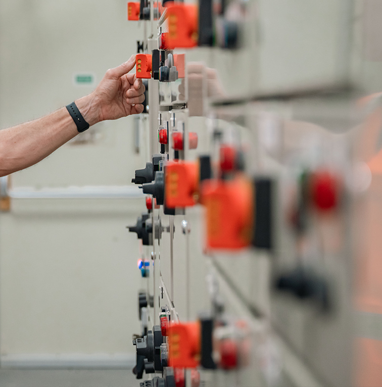 A worker in a high-visibility vest adjusts a switch on an electrical control panel filled with buttons, switches, and indicator lights.