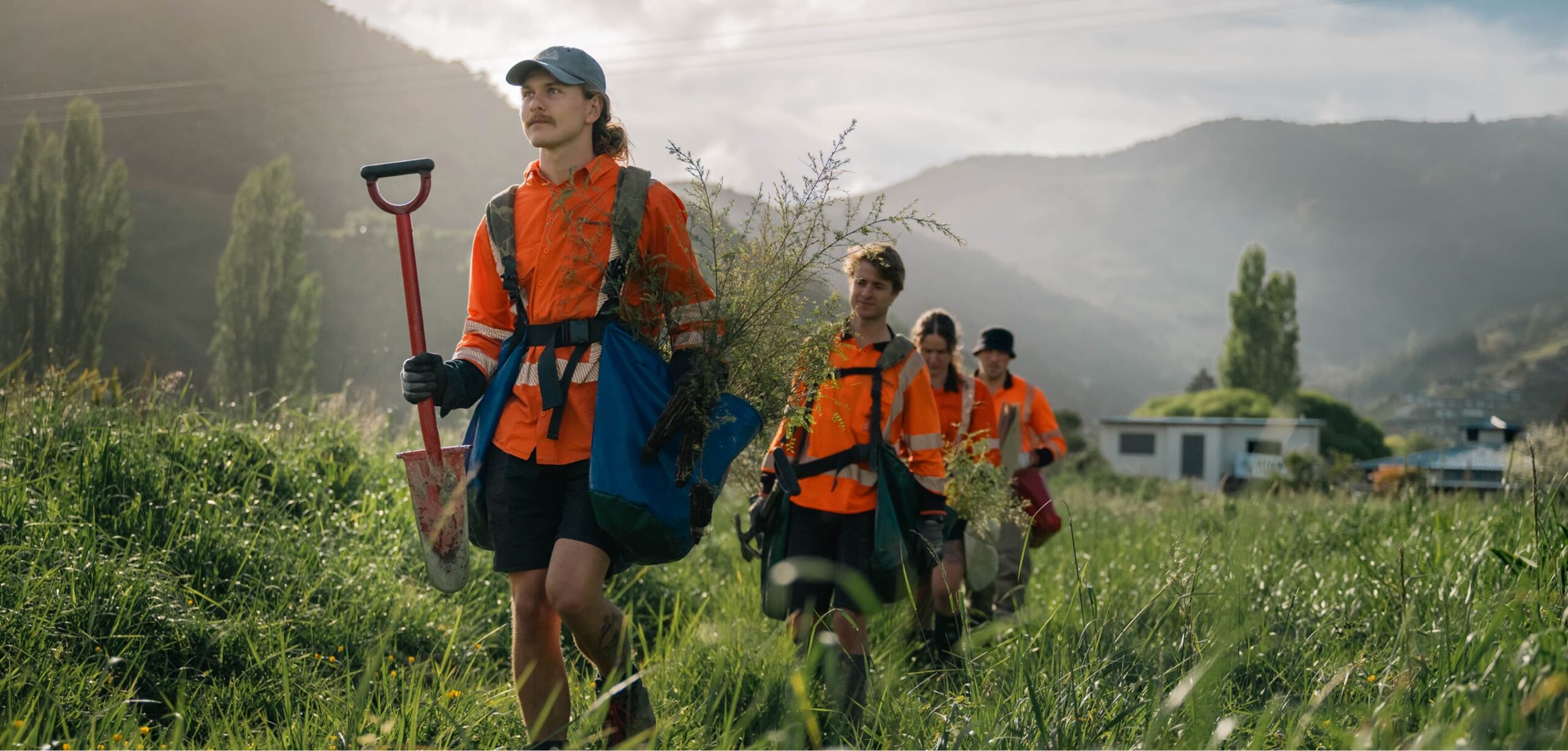 A group of Kūmānu field workers in high-visibility gear walk through tall grass carrying tools and native seedlings, with misty hills in the background.