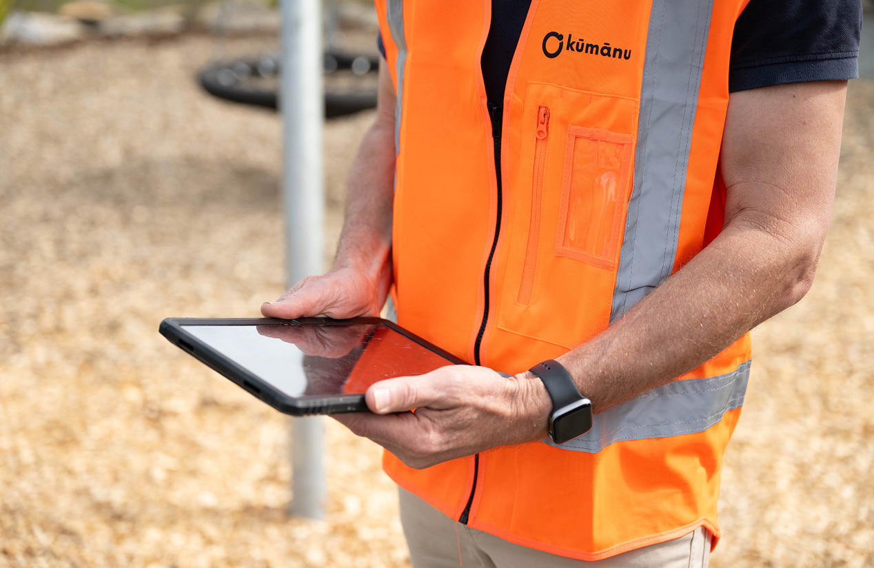 Close-up of a worker in an orange hi-vis vest holding a tablet outdoors, with the Kūmānu logo visible on their uniform.