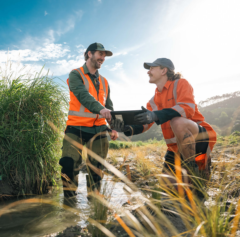Two Kūmānu team members in high-visibility gear stand in a shallow stream reviewing information on a tablet while carrying out fieldwork.