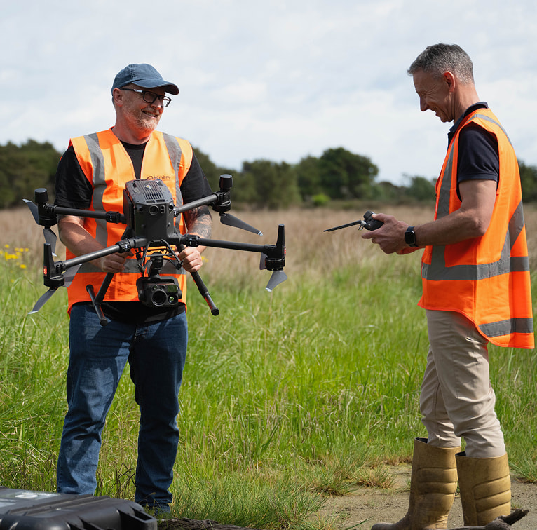 Two Kūmānu team members in high-visibility vests prepare a large field drone for survey work in an open grassland area.