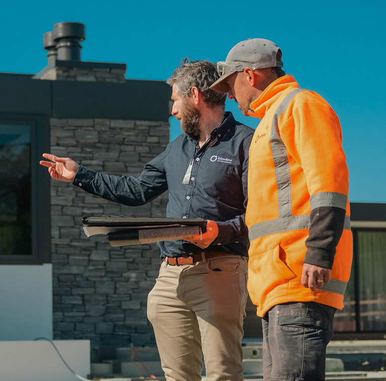 Two Kūmānu team members on a landscape worksite reviewing plans outdoors, with one person gesturing toward the project area.