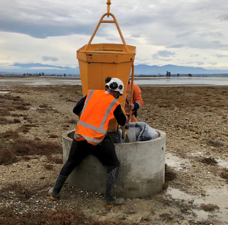 Two workers in orange safety vests installing a concrete structure in a coastal, rocky landscape with distant mountains and overcast skies.