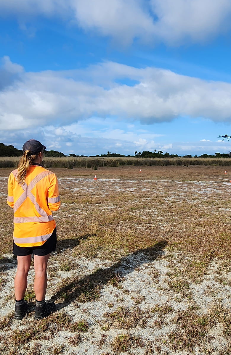A woman wearing a high-visibility orange shirt stands in an open field while operating a drone flying ahead of her under a partly cloudy sky.