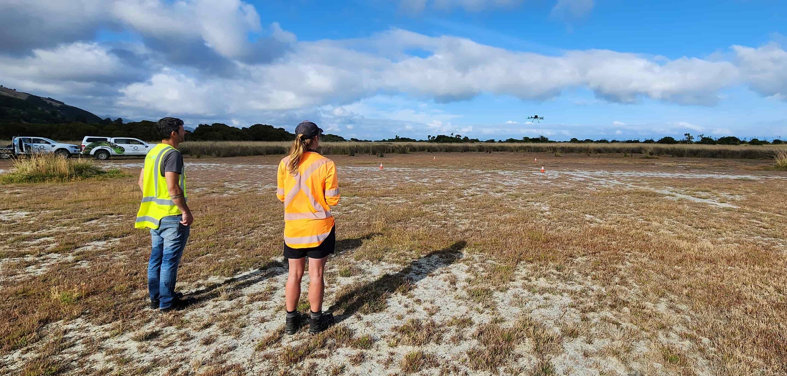 Two people wearing high-visibility vests stand in an open, dry grass field while watching a drone flying in the distance, with work vehicles parked behind them and a blue sky overhead.