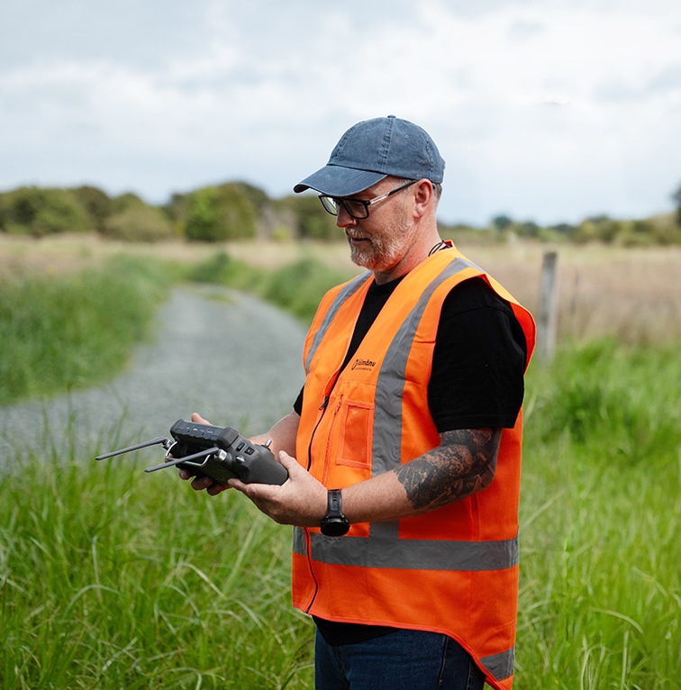 A man in a high-visibility orange vest and cap holds a drone controller while standing on a grassy track, preparing to operate a drone.