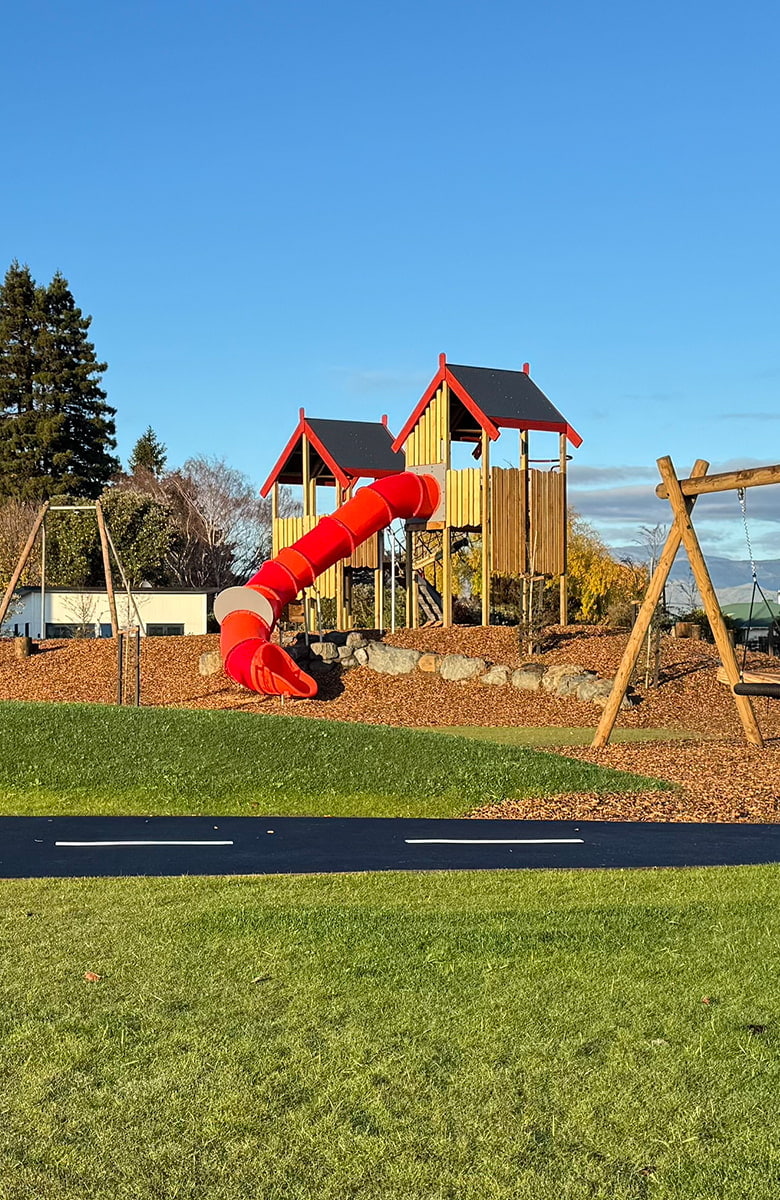 A close-up view of a bright red tube slide attached to wooden play structures in a landscaped playground