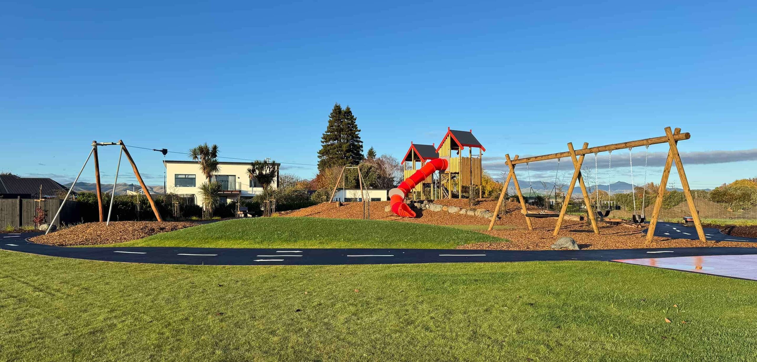 A modern playground featuring swings, a zipline, and a large red tube slide, set within a landscaped park under a clear blue sky