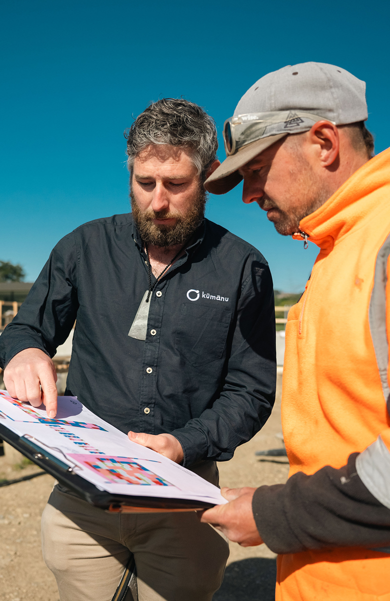 A Kūmānu team member reviews colourful project plans with a worker in high-visibility gear on an active construction site under a clear blue sky.
