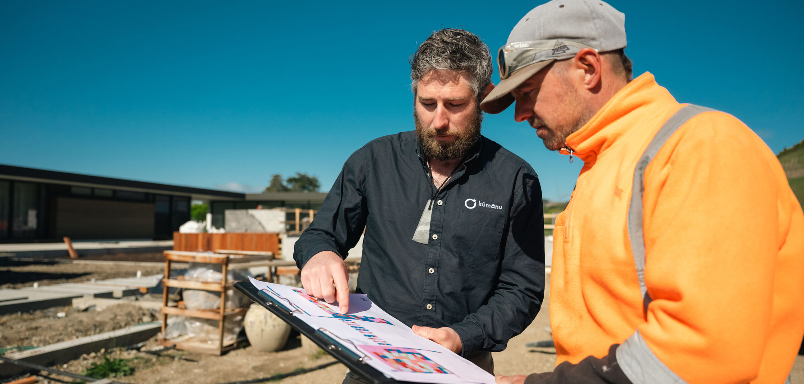 A Kūmānu team member reviews colourful project plans with a worker in high-visibility gear on an active construction site under a clear blue sky.