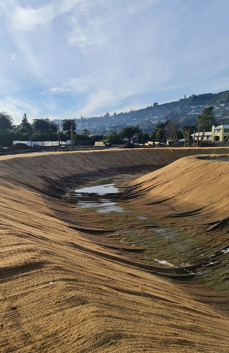 A large, newly formed stormwater or wetland basin lined with erosion-control matting at an active construction site, with machinery, materials, and surrounding hills in the background.