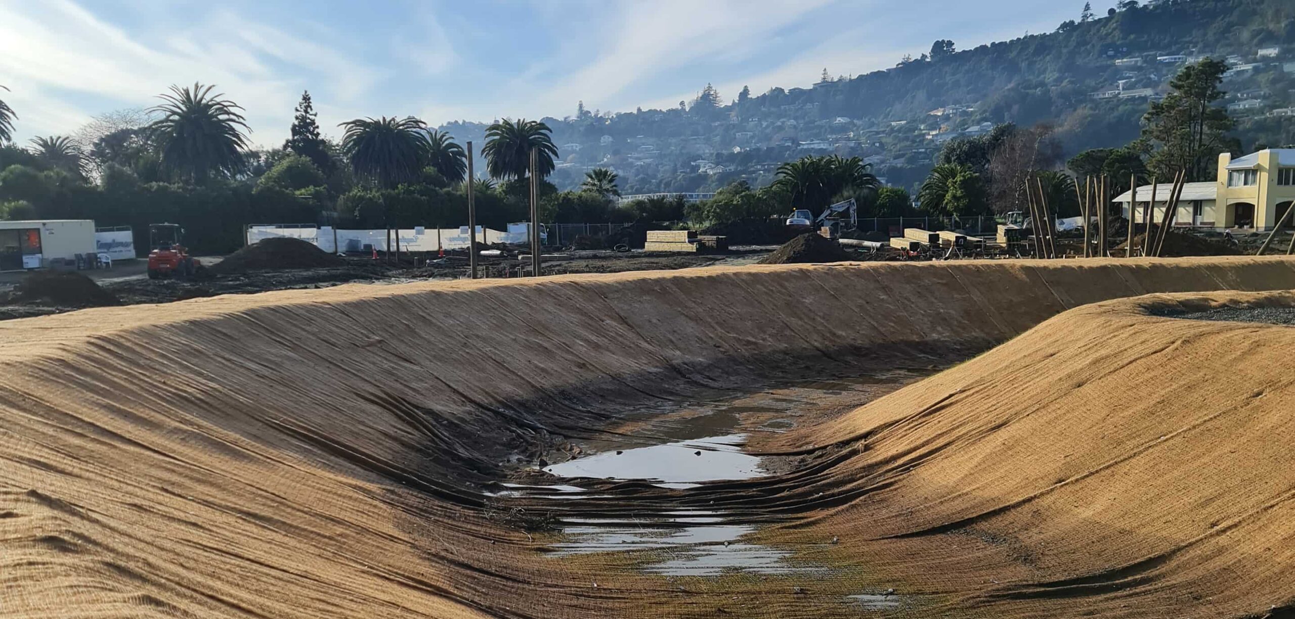 A large, newly formed stormwater or wetland basin lined with erosion-control matting at an active construction site, with machinery, materials, and surrounding hills in the background.