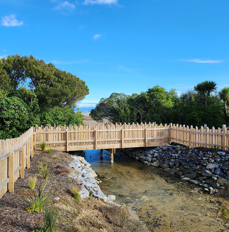 A wooden pedestrian bridge with fencing spans a shallow stream, surrounded by native vegetation and landscaped plantings under a bright blue sky.