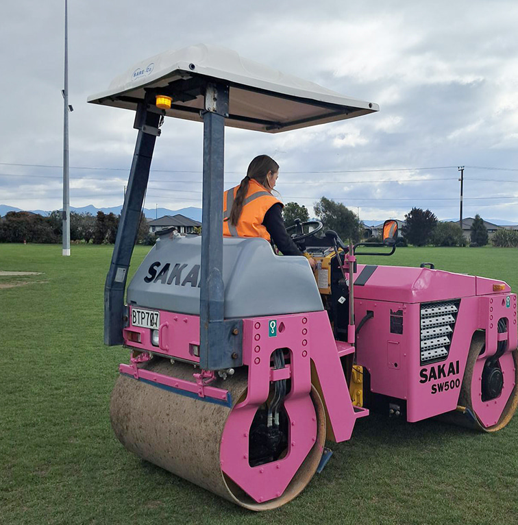 A Kūmānu team member in high-visibility gear operates a pink turf roller across a large grass field on an overcast day