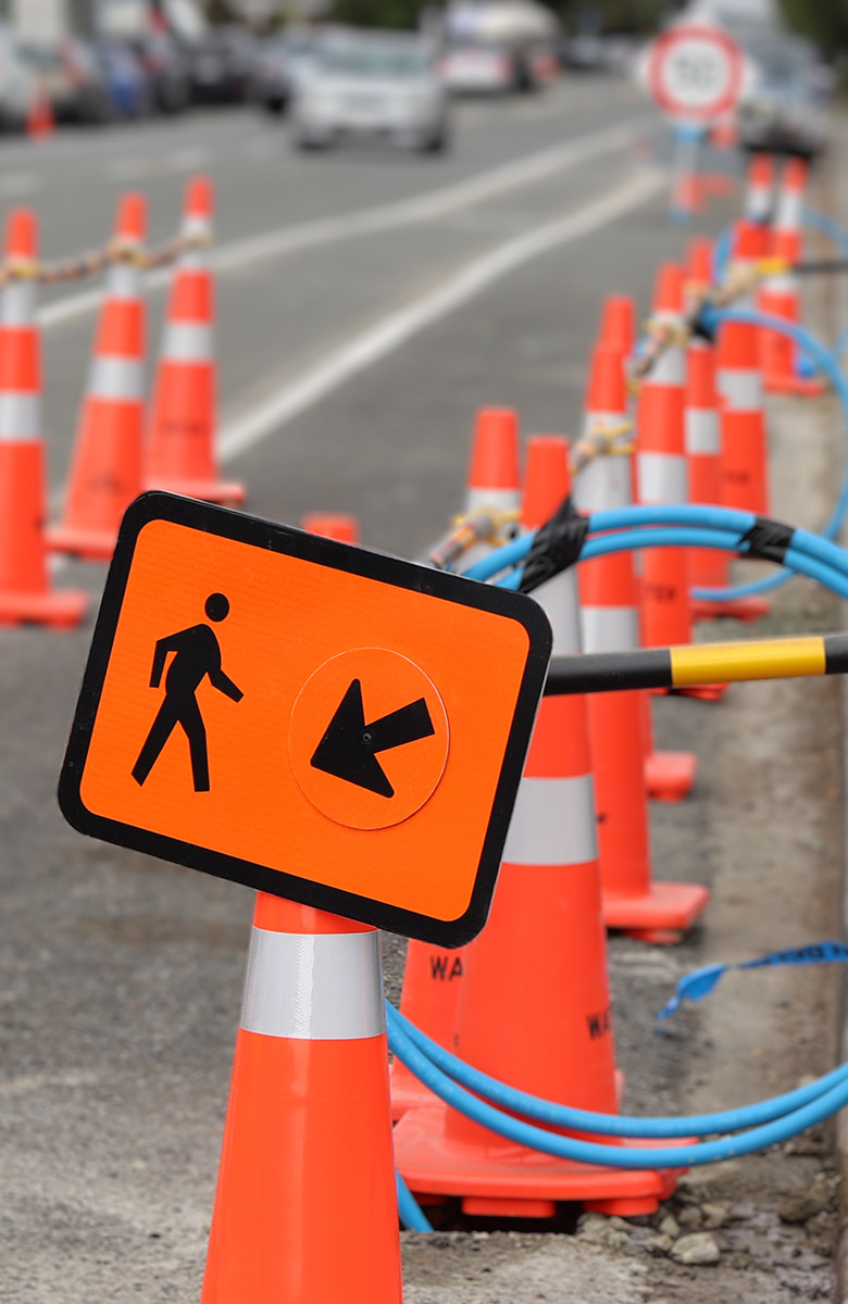 Close-up of a pedestrian detour sign mounted on an orange cone, surrounded by orange traffic cones and blue cabling along a roadside construction zone