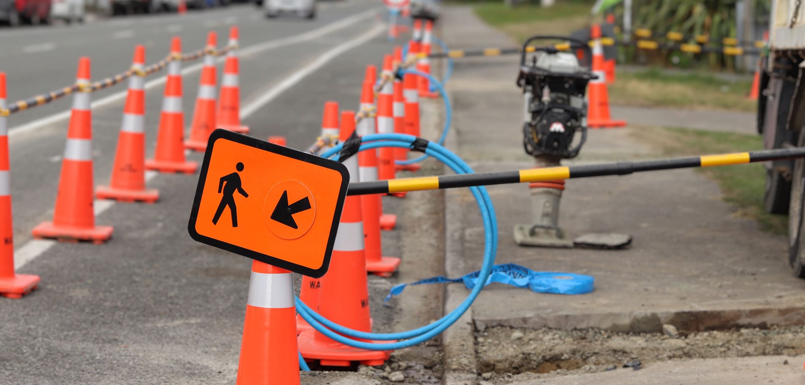 Roadwork scene with rows of bright orange traffic cones lining a city street, a pedestrian detour sign pointing left, and construction equipment visible in the background
