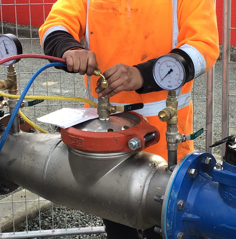 Worker in a high-visibility orange jacket adjusting gauges and valves on a large stainless-steel water pipe fitted with pressure meters and testing equipment.