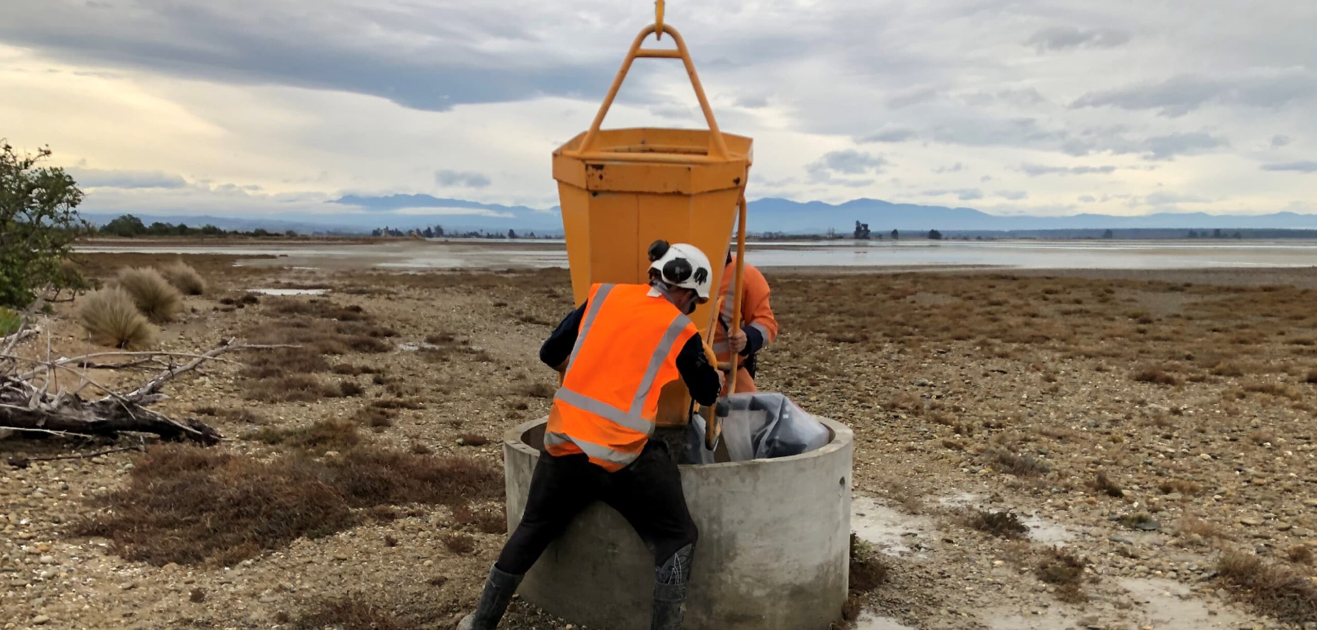 Workers in high-visibility gear pour material into a concrete manhole structure on a coastal construction site