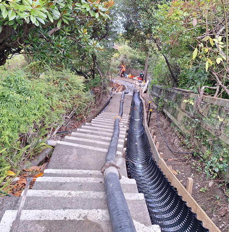 A steep outdoor staircase under construction, with drainage pipes and materials laid along the steps in a bush-covered area