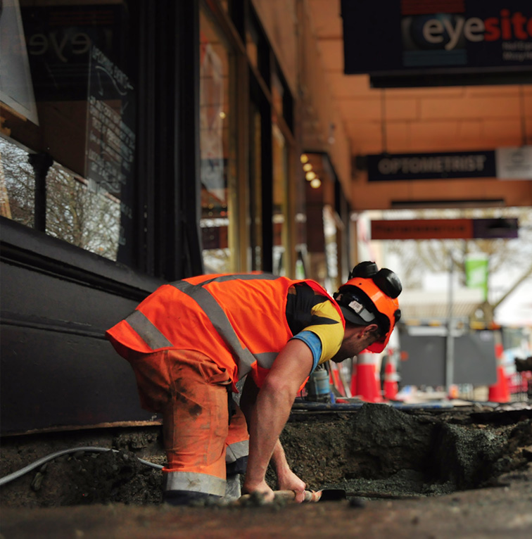 Worker in high-visibility orange gear kneeling to repair a footpath in a town center, with shops and construction cones in the background.