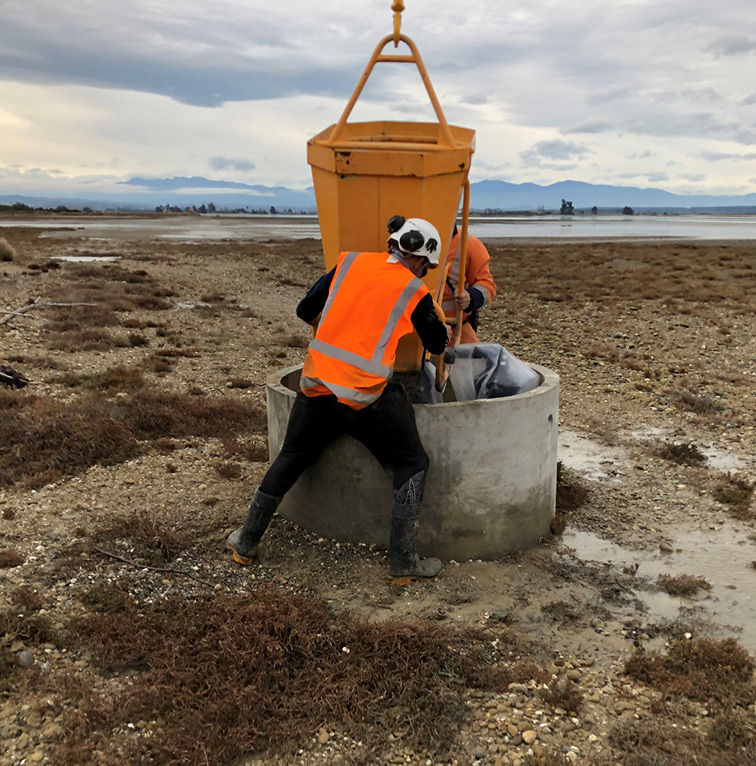 Two workers in orange safety vests installing a concrete structure in a coastal, rocky landscape with distant mountains and overcast skies.