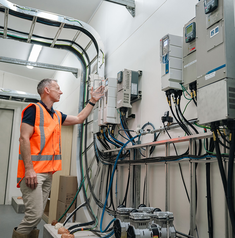 Technician wearing a high-visibility vest inspecting a wall of electrical control panels inside an industrial facility.