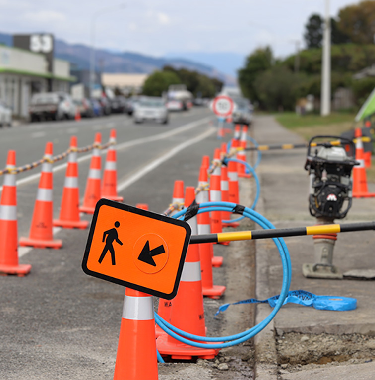 Roadside lined with orange traffic cones and a pedestrian detour sign, with vehicles and shops visible further down the street.