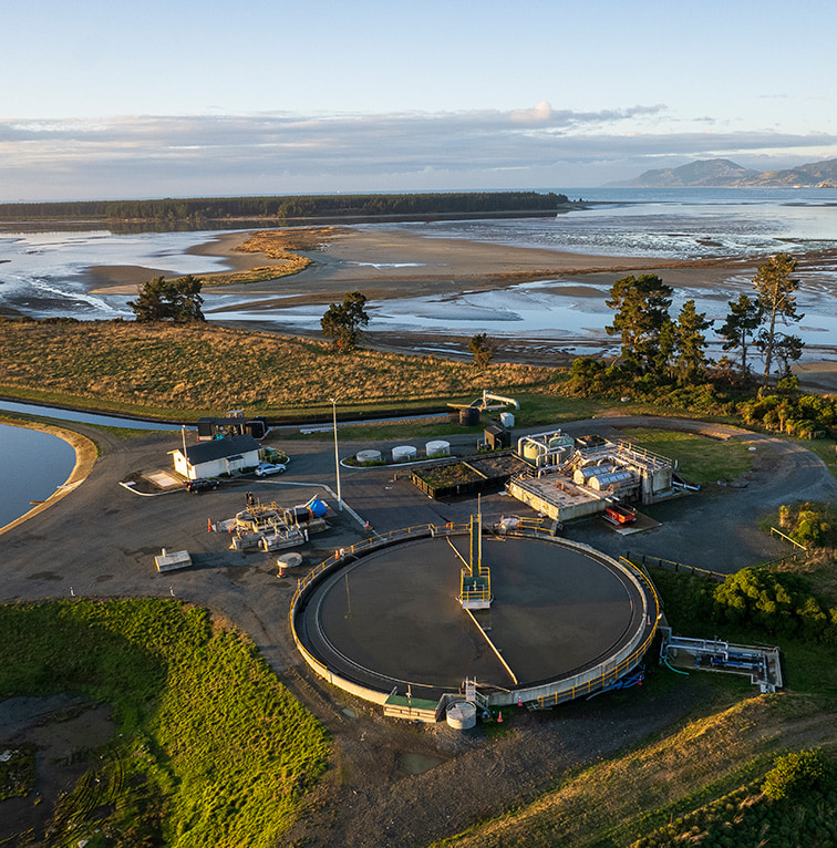 Aerial view of a water treatment facility beside a coastal estuary, with ponds, tanks, and surrounding landscape at sunrise.