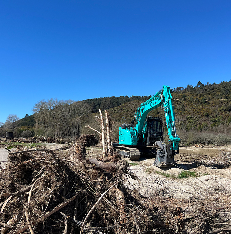 A teal excavator clearing and moving large piles of uprooted trees and debris on a sunny day, with hills and bush in the background.