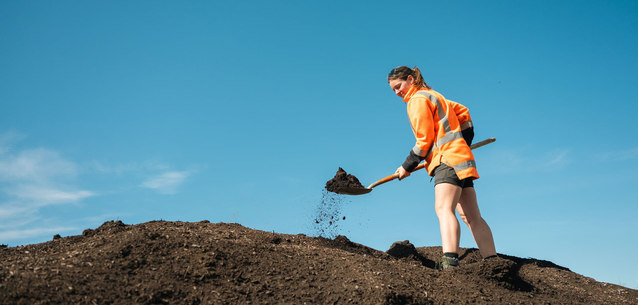 Worker in a high-visibility orange jacket shovelling a pile of soil on a sunny day, with a clear blue sky in the background.