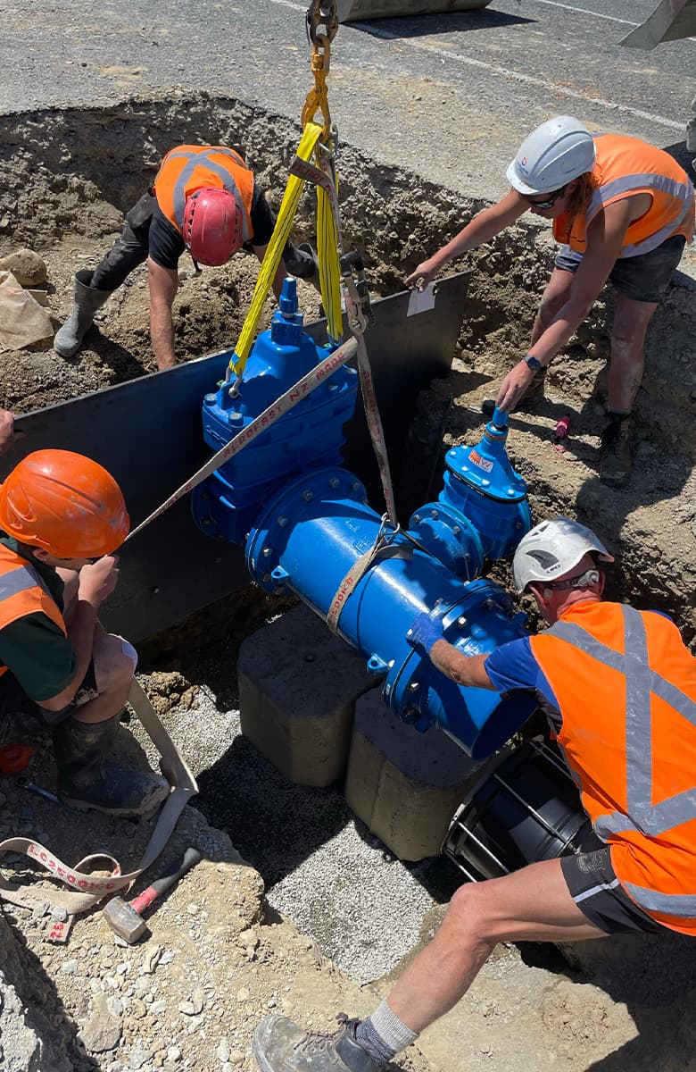 Construction team positioning a large blue water valve into place using lifting equipment inside a trench