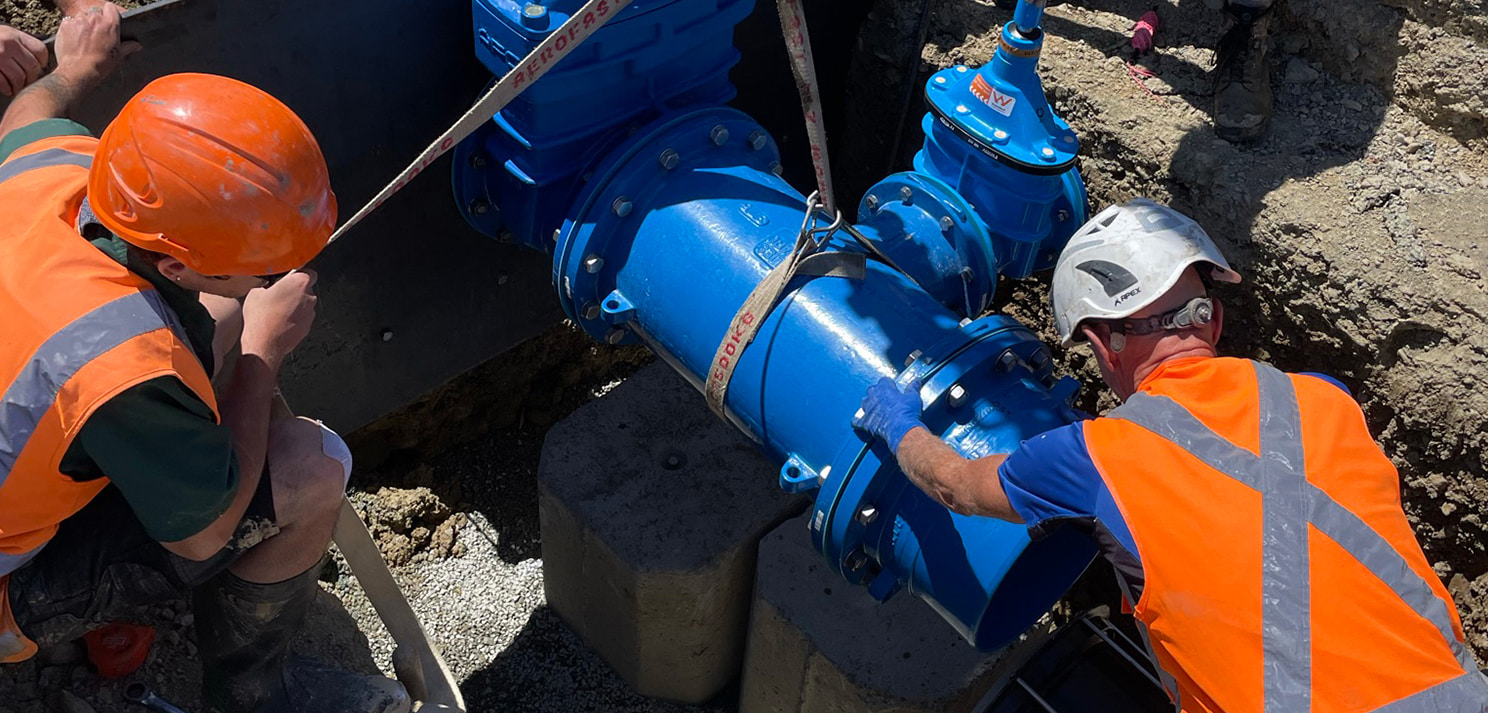 Workers in high-visibility clothing installing a large blue water pipeline valve in a trench using lifting straps