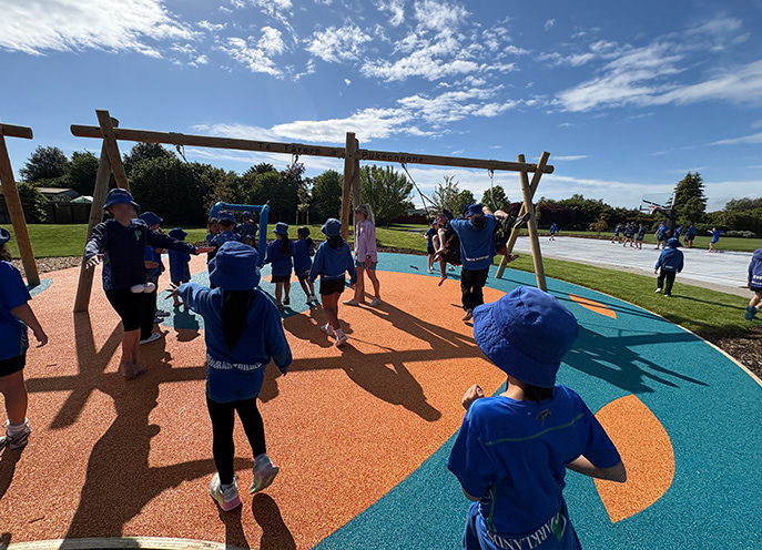 Group of children in blue uniforms playing around a timber swing set on blue and orange rubber surfacing at Decks Reserve Playspace