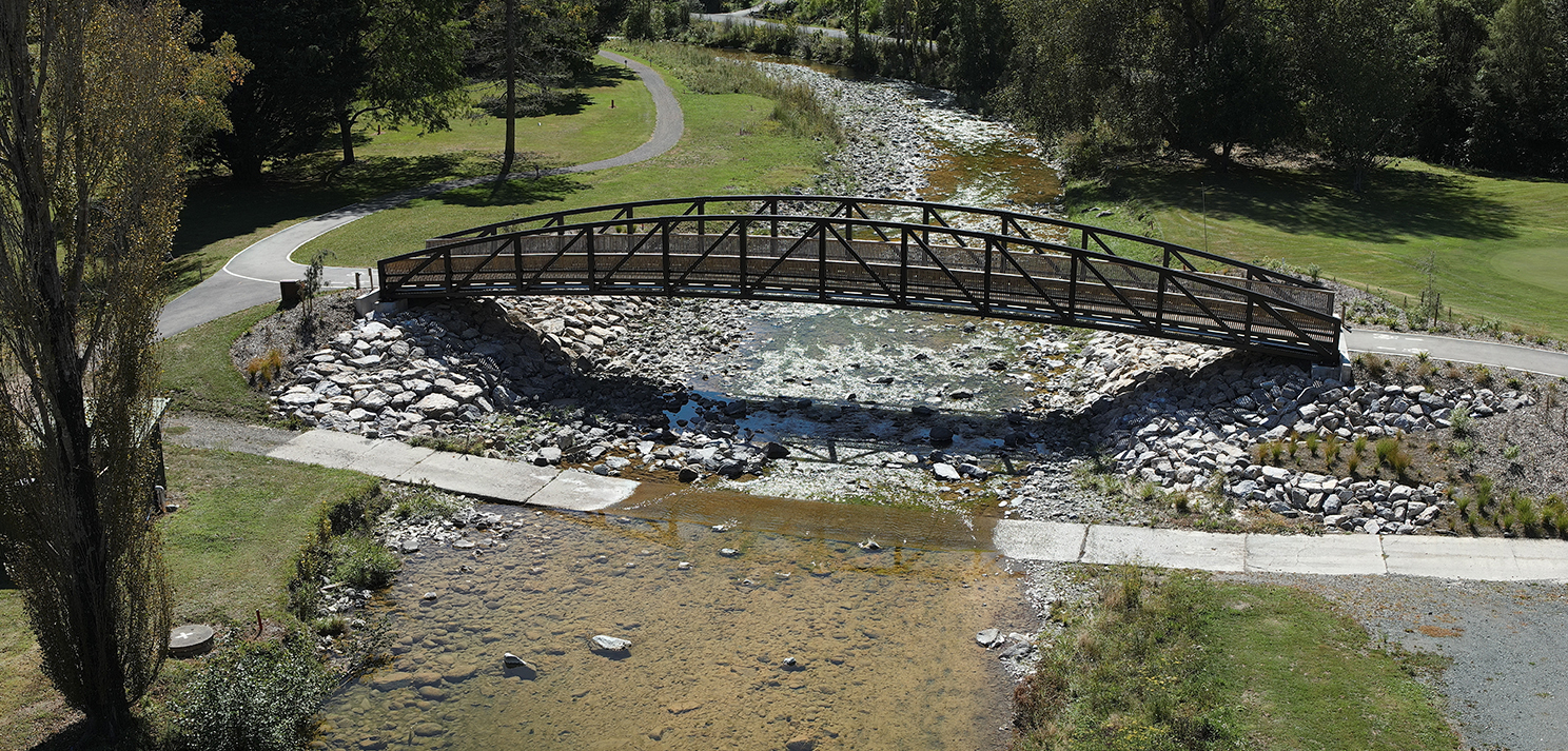 Timber pedestrian bridge spanning a shallow stream with rock-lined banks and surrounding parkland