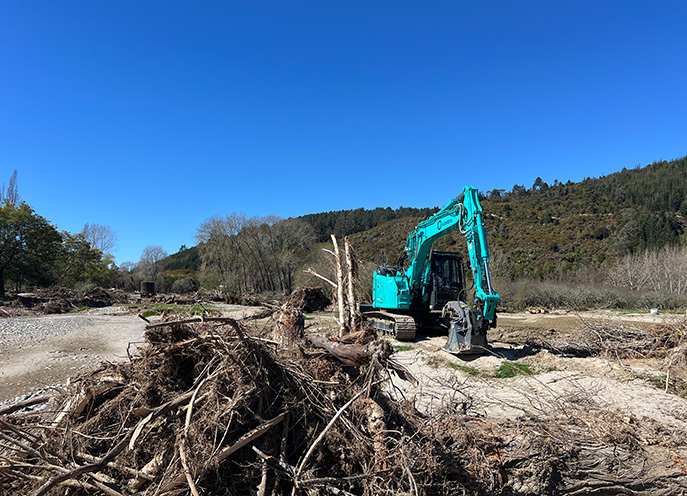 Excavator working beside large piles of flood debris and exposed riverbed sediments