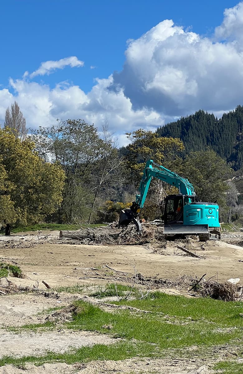 Portrait-oriented view of an excavator removing flood debris from a riverbed with trees and hillside beyond