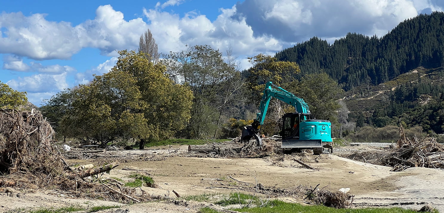 Excavator clearing flood debris and sediment from a riverbed with trees and hills in the background