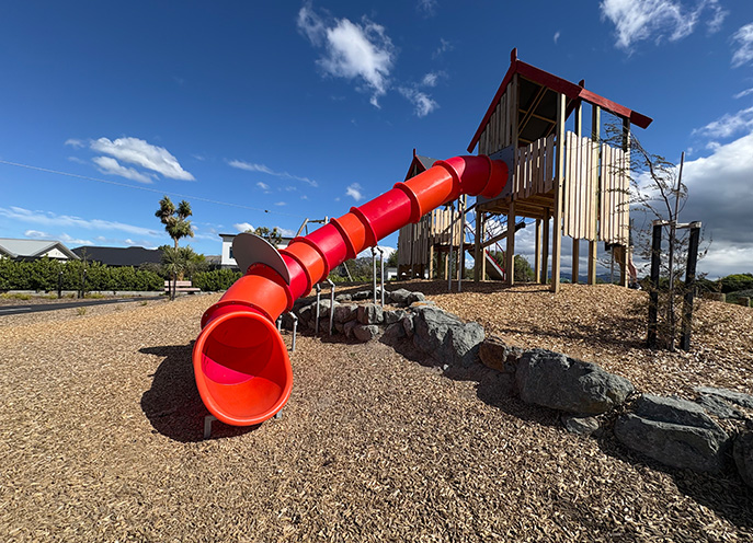 Red tube slide attached to timber play towers on a raised bark-covered mound