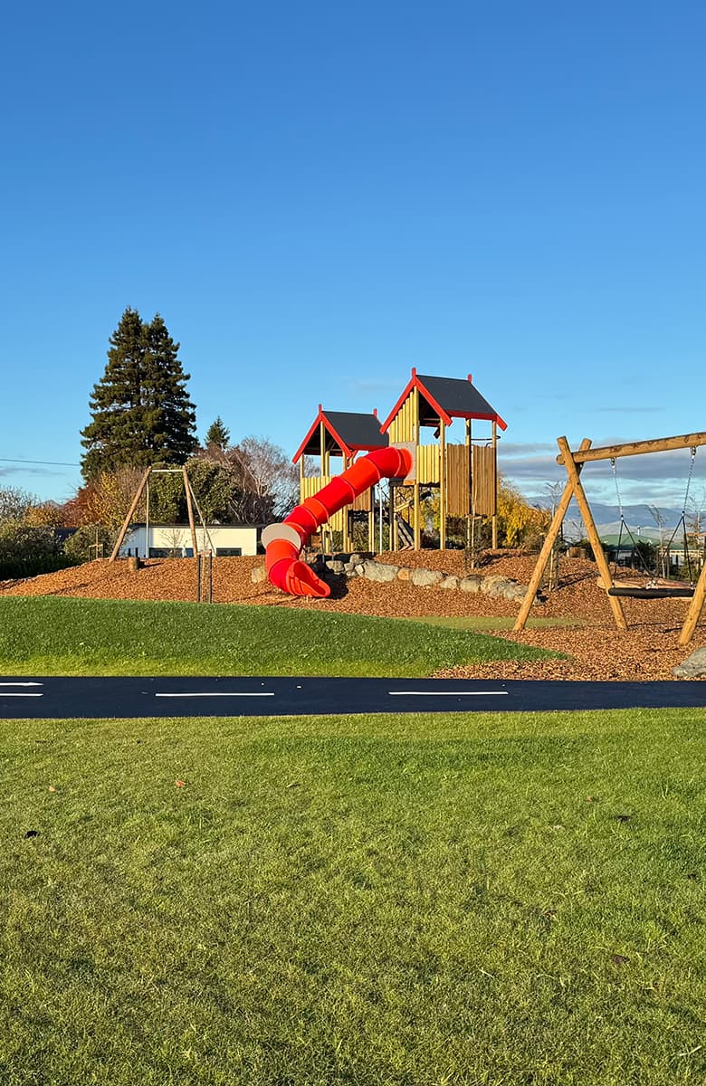 Portrait-oriented view of Rose Manor Playspace with play towers, red tube slide, swings, and grassy foreground