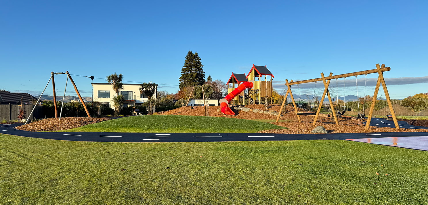 Wide view of Rose Manor Playspace showing swings, play towers with red tube slide, grass areas, and surrounding houses
