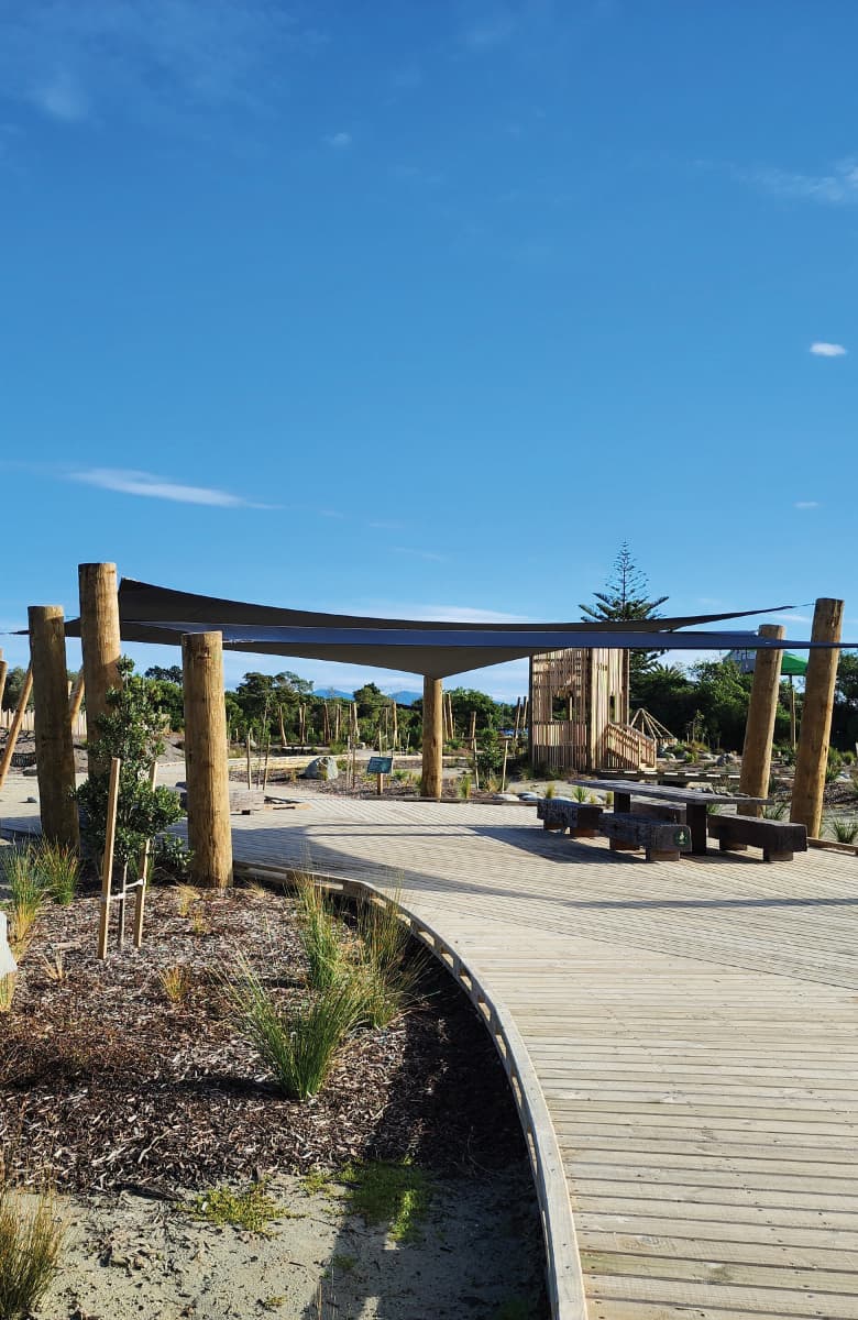 Curved timber boardwalk with shade sail and seating within the Te Pā Harakeke community playspace